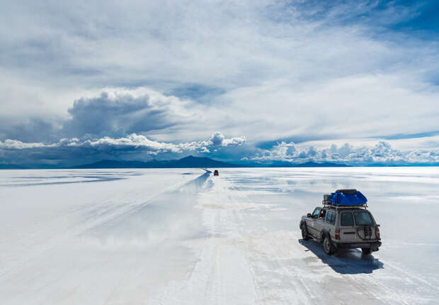 An SUV drives across a vast, reflective salt flat under a dramatic, cloud-filled sky, with mountains visible in the distance.