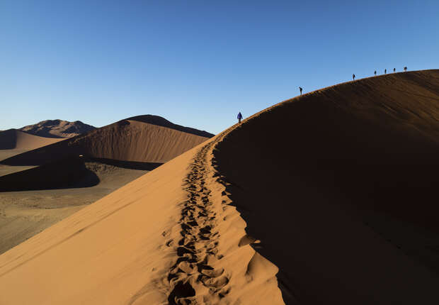 Footprints trail along a steep sand dune as several people walk upwards under a clear blue sky, casting long shadows in the desert landscape.