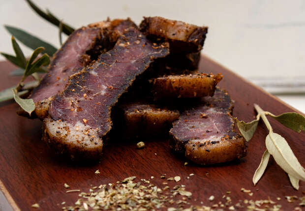 Slices of cured, seasoned meat are stacked on a wooden board. Surrounding the meat are fresh herbs and scattered spices, set against a light, neutral background.