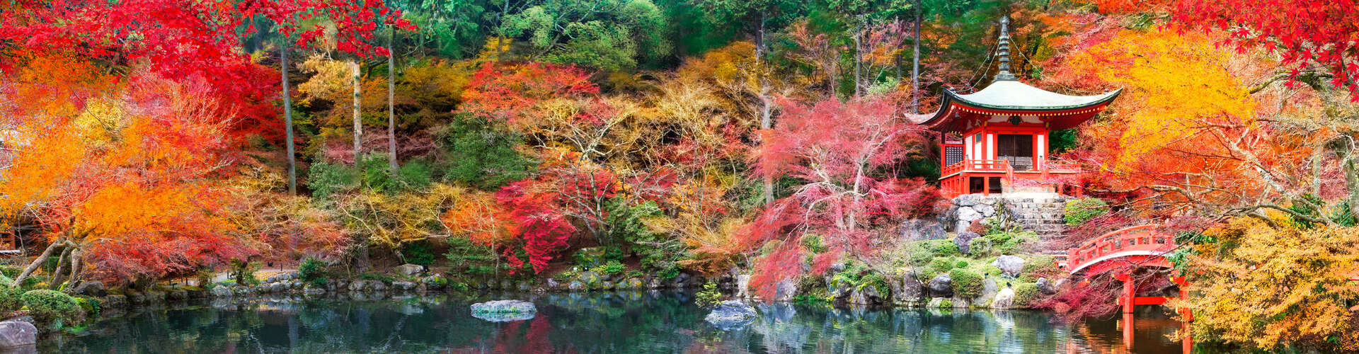 Pagoda with red and white detailing sits beside a curved red bridge, surrounded by vibrant autumn foliage in orange, yellow, and red near a tranquil reflective pond.