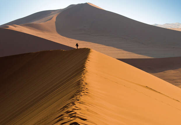 A lone person walks along a high, narrow sand dune ridge in a desert, with expansive orange dunes and clear blue sky surrounding them.