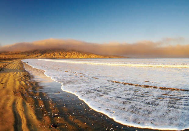 Waves gently wash onto a sandy beach under a clear blue sky, with distant fog obscuring low, rolling hills on the horizon.