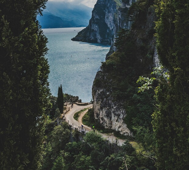 A winding path curves along a cliffside, bordered by lush greenery, overlooking a serene blue lake under a misty, mountainous horizon.