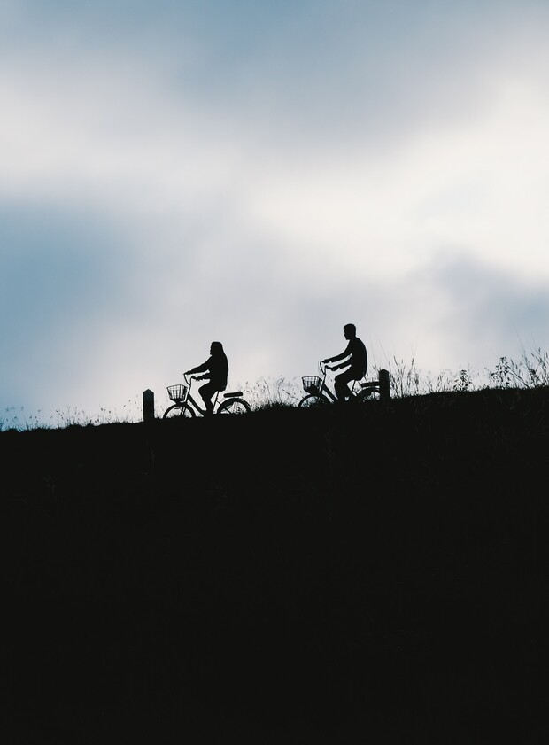 Silhouetted cyclists ride along a grassy hill against a cloudy sky, creating a serene outdoor scene.