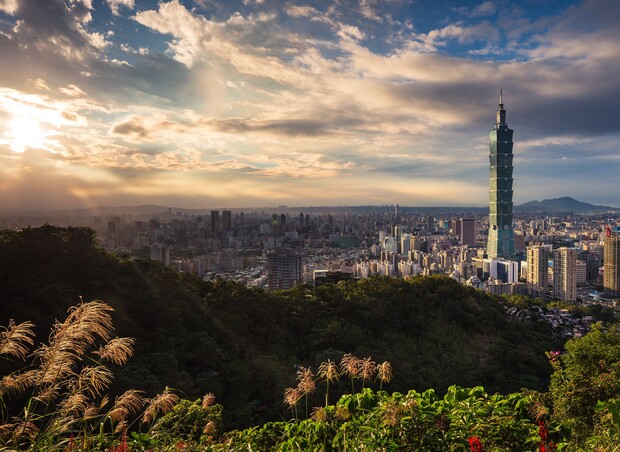A tall skyscraper stands prominently against a vibrant sunset sky, surrounded by an expansive cityscape and lush greenery in the foreground.
