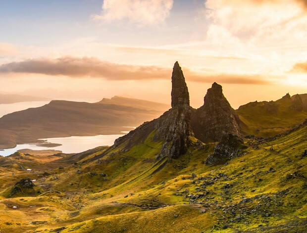 Two jagged rock formations tower over a green, hilly landscape at sunrise, with a serene lake and distant mountains under a partly cloudy sky.