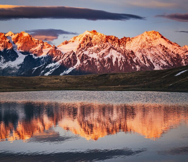 Snow-capped mountains reflect in a calm lake at sunset, displaying vivid orange and pink hues. Surrounding grassy terrain blends into the serene landscape, under a partly cloudy sky.