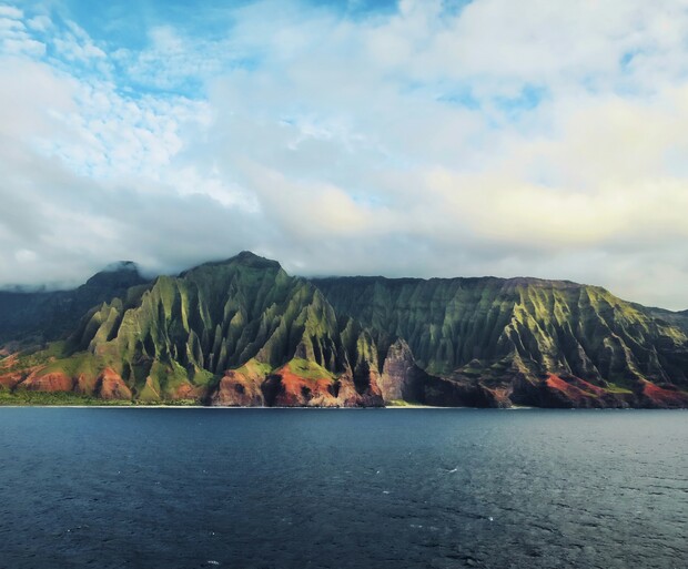Cliffs rise majestically, covered in lush greenery and red hues, standing against a backdrop of a cloudy sky. The calm ocean stretches in the foreground, enhancing the serene landscape.
