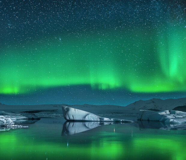 Northern lights shimmer across a starry night sky, illuminating icebergs floating in tranquil water with a mountainous horizon in the background.