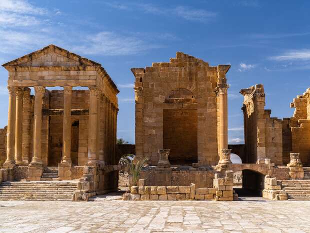 Ancient stone ruins stand with partially-intact columns and archways on a worn, cobbled surface under a clear blue sky, conveying historical significance and architectural grandeur.