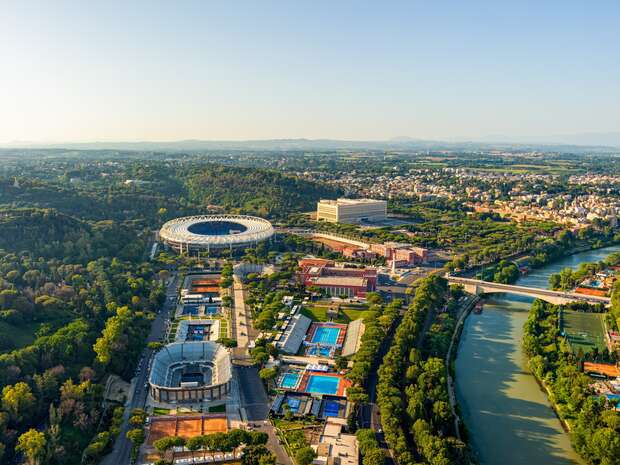 Aerial view of a large stadium nestled in lush greenery with adjacent sports facilities and a river flowing alongside, bordered by a bridge, with a cityscape in the background.