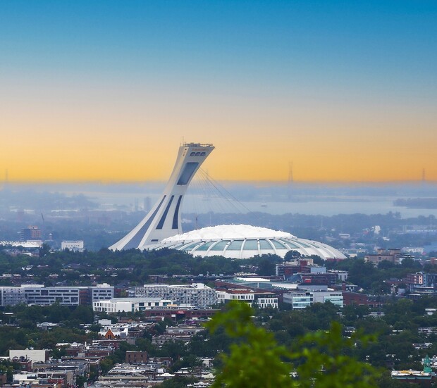 A large, white stadium with a distinctive leaning tower rises above a cityscape, surrounded by buildings and greenery, under a colorful sky transitioning from blue to orange at sunset.