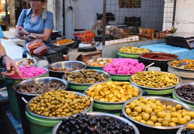 Bowls of various pickled foods, including olives and pink turnips, are displayed in a bustling market, with people browsing nearby amidst a vibrant background of other market goods.