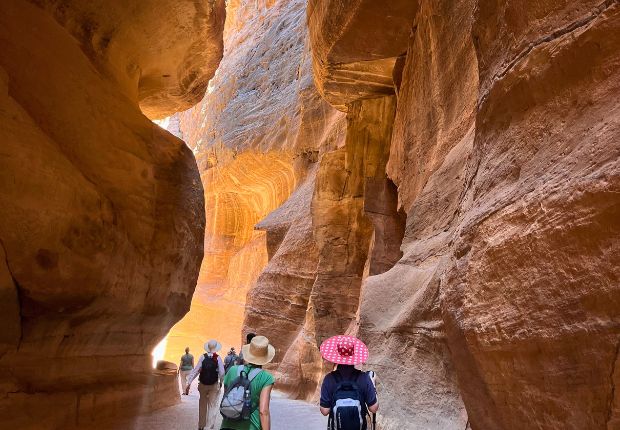 Hikers walk through a narrow canyon with towering sandstone walls, lit by sunlight filtering from above. They wear hats and backpacks, exploring the natural passageway.