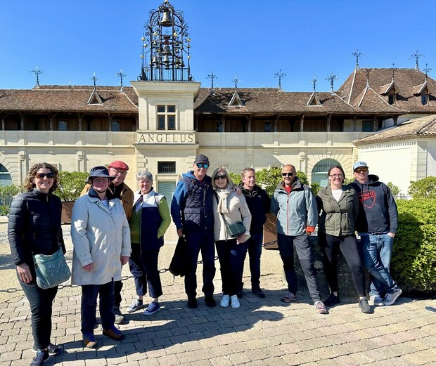 A group of ten people stands on a cobblestone path, smiling for the camera. Behind them is a large building with a sign reading "ANGELUS," adorned with a decorative roof and spire.