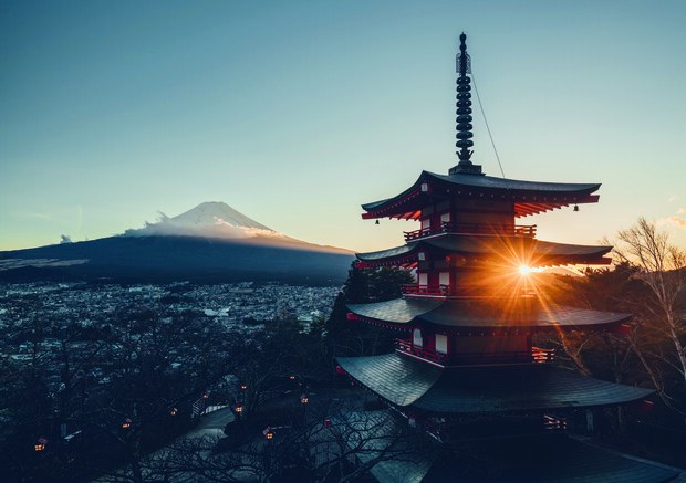 A pagoda catches the sunrise, its silhouette contrasted against Mount Fuji in the background, surrounded by a quiet landscape of trees and distant cityscape.