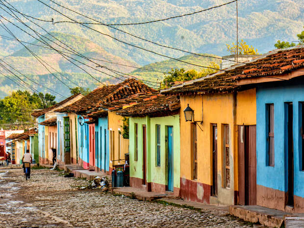 Colorful houses line a cobblestone street, while a person walks away. Overhead wires stretch across the scene, set against a backdrop of lush, mountainous terrain.