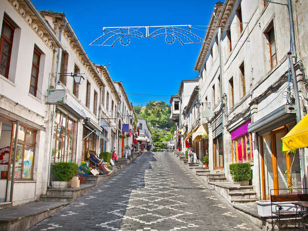 A cobblestone street ascends between rows of historic buildings with colorful awnings, leading to greenery under a clear blue sky; overhead, a decorative metal framework spans the street.
