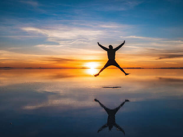 A person jumps in the air with arms and legs outstretched against a vibrant sunset over a reflective, water-covered surface, creating a striking mirrored silhouette.