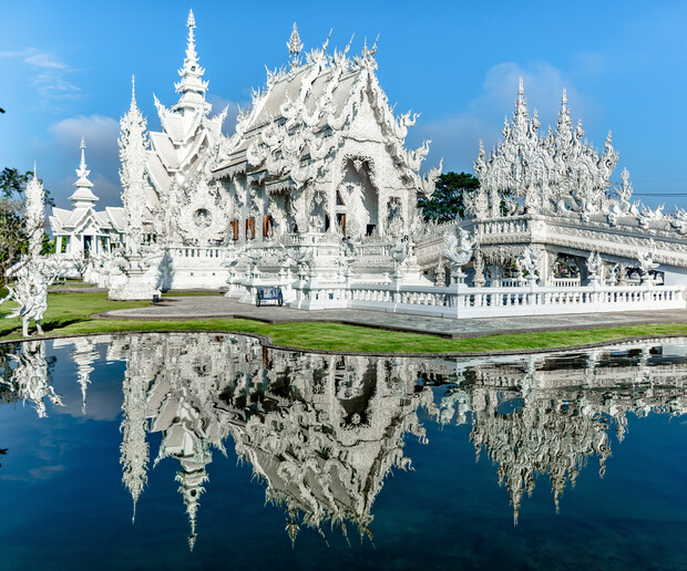 An intricate white temple is reflected in a calm pond. Surrounded by manicured grass and under a clear blue sky, its ornate design features spires and statues.