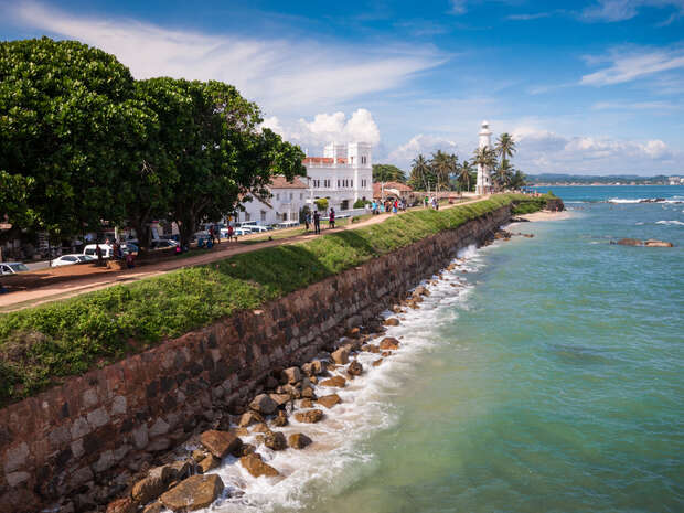 A lighthouse stands on a coastal path, bordered by trees and a historic fort wall. People walk nearby, overlooking the ocean under a partly cloudy blue sky.