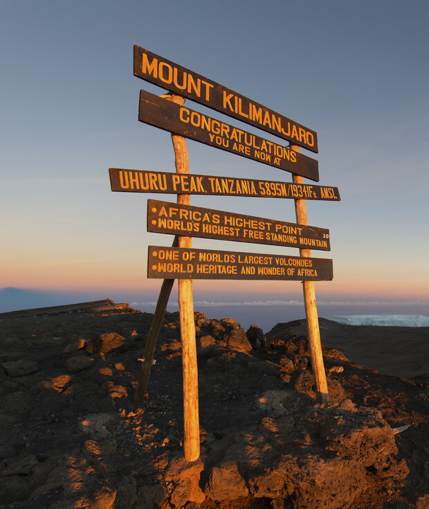 Wooden sign with multiple plaques reads "Mount Kilimanjaro, Congratulations, You are now at Uhuru Peak, Tanzania 5895m/19341ft AMSL. Africa's highest point, World's highest free standing mountain, One of world's largest volcanoes, World heritage and wonde