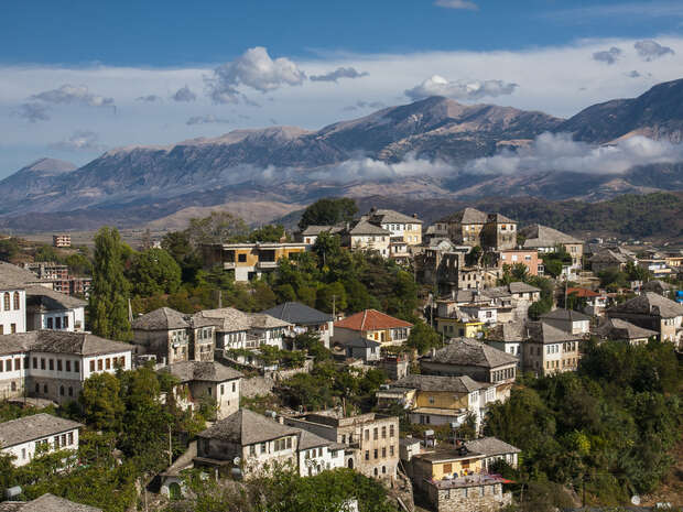 Stone-roofed buildings sit clustered on a hillside, surrounded by lush greenery. In the background, misty mountains rise under a cloudy sky. The scene is serene and picturesque.