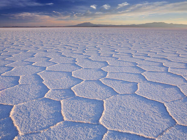 Salt flats with hexagonal patterns stretch across the foreground, under a vast sky with clouds. Distant mountains are visible on the horizon, bathed in soft sunlight.