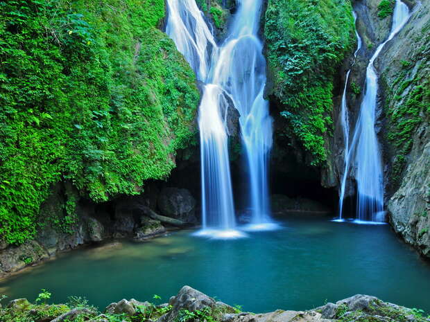 Waterfalls cascade into a clear blue pool, surrounded by lush green foliage and rocky cliffs, creating a serene and natural setting.