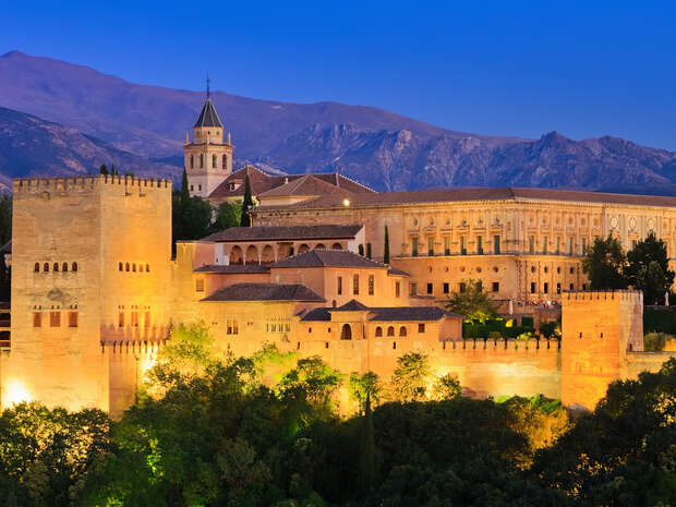 Ancient fortress illuminated at dusk, highlighting its detailed architecture. Surrounded by lush greenery, the structure stands against a backdrop of mountains and a clear, deep blue sky.