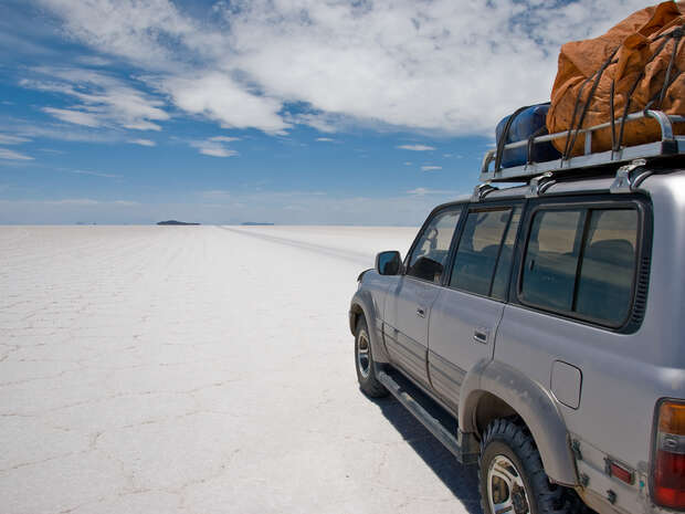 SUV parked on vast, cracked salt flat under a partly cloudy sky, with luggage secured on the roof, extending to the horizon.