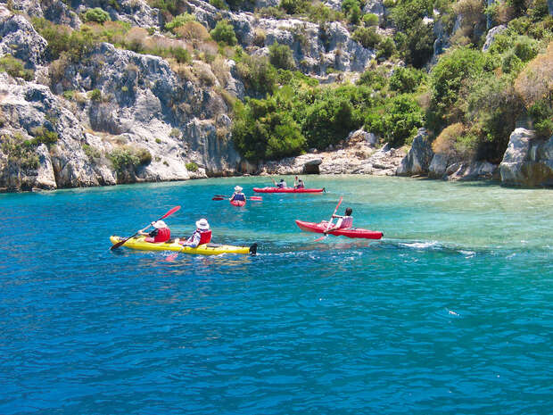 Kayaks glide across vibrant blue water, paddled by individuals in red life vests, set against a rocky shoreline with lush greenery under a clear sky.