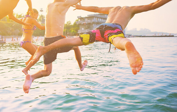 Three people jump into a body of water with lively expressions, wearing swimwear. They are mid-air against a sunny backdrop, while buildings and greenery line the distant shore.