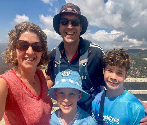A smiling group of four people stands closely together outdoors, wearing casual attire and hats, with a cloudy sky and mountainous landscape in the background. They're enjoying a sunny day.