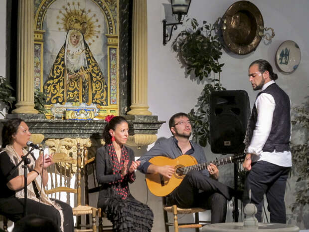 A guitarist plays while three people, including a standing man, engage in a performance. A decorated religious painting and potted plants adorn the background, creating an intimate cultural setting.