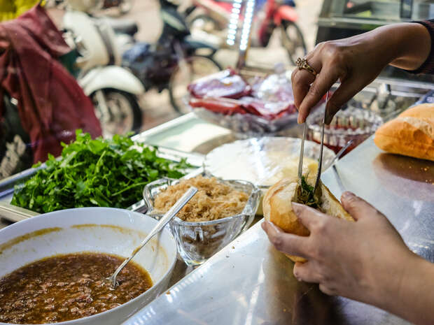 Hands preparing a sandwich by filling a baguette with ingredients using tongs. Nearby, fresh greens and sauces are arranged on a counter. Motorbikes are visible in the street outside.