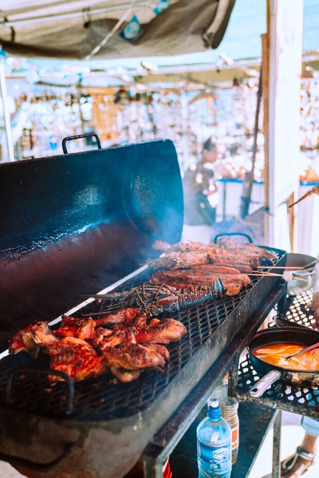 Grilled meats cooking on an open barbecue, producing smoke, in an outdoor market setting with various stalls visible in the background. Bottles and a pan with sauce are nearby.