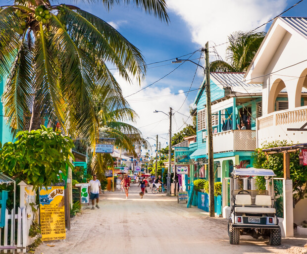 A golf cart is parked on a sandy street lined with colorful buildings and palm trees, people walking nearby. Signs say "Friends of Belize" and "Coffee in the House."