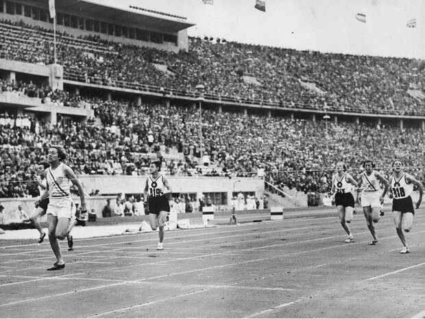 Athletes sprint on a track, with one leading. They wear numbered vests (118, 88, 118). A packed stadium with spectators and flags forms the backdrop.