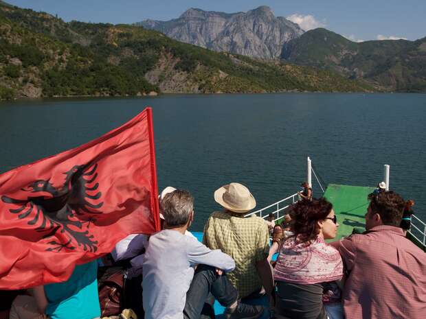 People sit on a boat deck adorned with an Albanian flag, gazing at a scenic lake surrounded by lush mountains under a clear blue sky.