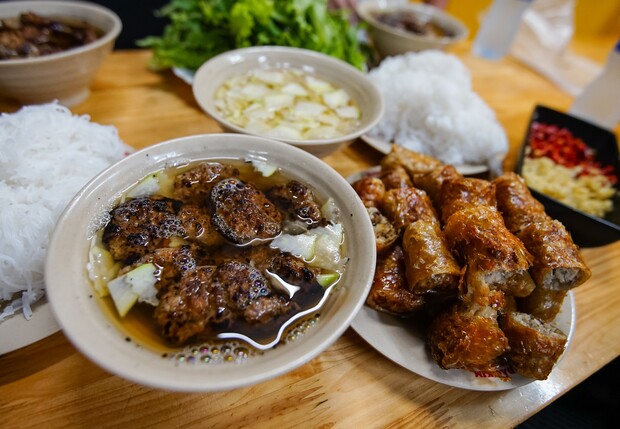 A bowl of grilled meatballs floats in broth, surrounded by plates of spring rolls, vermicelli noodles, and fresh greens, set on a wooden table in a dining setting.