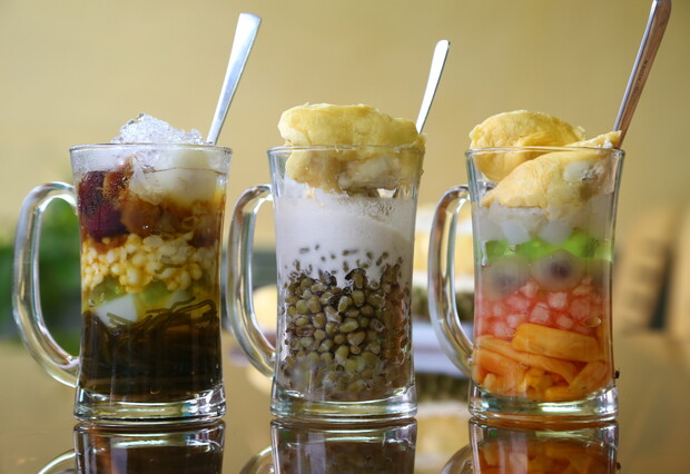 Three glass mugs hold colorful, layered desserts with spoons. Each dessert features shaved ice, various beans, fruits, and a scoop of durian, in a dining setting with a smooth backdrop.