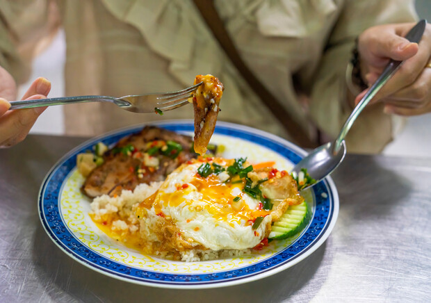 A person holds a forkful of grilled meat with rice and a fried egg, drizzled with chili sauce, on a decorative blue and yellow plate at a dining table.