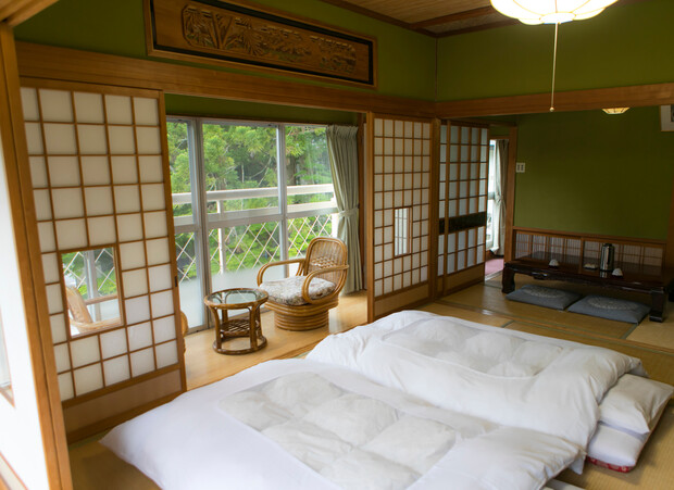 Futons lie neatly on tatami mats in a traditional Japanese room, featuring sliding shoji doors. A wicker chair and table are by a window, overlooking a green garden.