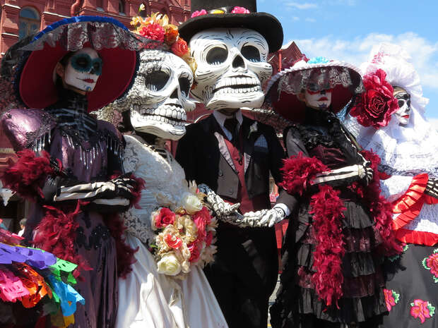 Figures dressed as skeletons wear elaborate, colorful costumes and large hats adorned with flowers, standing in a group during a festive outdoor event against a backdrop of red architecture and blue sky.