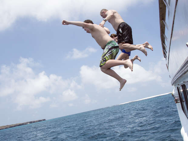 Three people jump together from a boat into the ocean, with clear blue skies and distant land visible in the background.