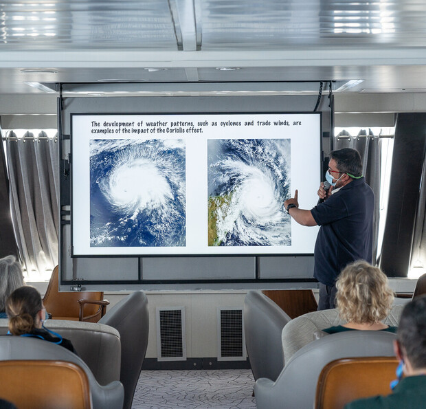 A presenter gestures towards a screen displaying satellite images of cyclones. The text reads, "The development of weather patterns, such as cyclones and trade winds, are examples of the impact of the Coriolis effect." Audience members are seated, watchin