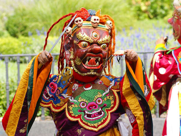 A performer in an elaborate, colorful mask and costume poses dramatically outdoors, surrounded by greenery and flowers, evoking a traditional or cultural festival scene.