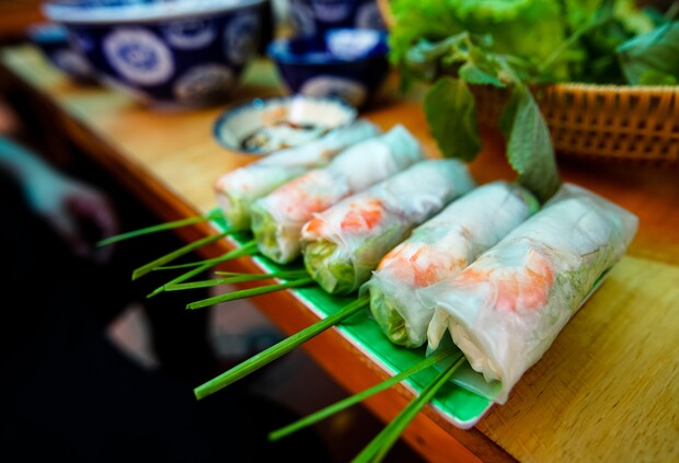 Fresh spring rolls with visible shrimp and herbs rest on a green plate. Nearby are blue bowls and a basket of leafy greens on a wooden table.