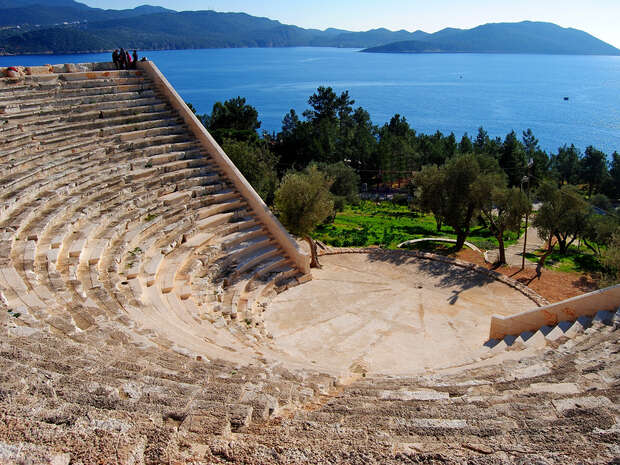 Semi-circular stone amphitheater curves downward, overlooking a grassy area with trees. Beyond, the scene opens to calm, blue sea and distant mountains under a clear sky.